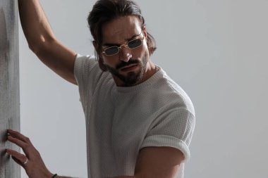 Güneş gözlüklü sert bir adam duvara dokunuyor ve gri arka planda kendine güvenen bir şekilde poz verirken gözünü kaçırıyor.