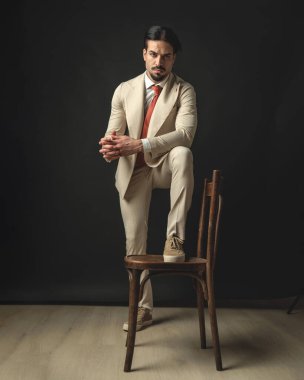 Bej takım elbiseli yakışıklı manken. Avuç içlerini bir arada tutarken ve dirseğini siyah arka planda tutarken sandalyenin üzerinde duruyor.