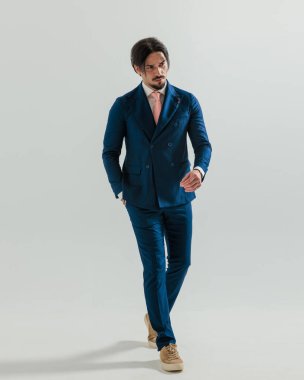 Donanma takımındaki moda adamı cebini tutarken öne doğru yürüyor ve gri arka planda yer almaya çalışıyor.