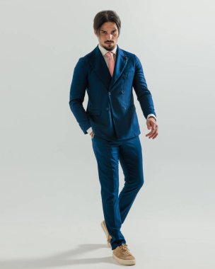 Çift göğüslü donanma takımlı bir adam gri arka planda cebini tutarken ileri doğru yürüyor.