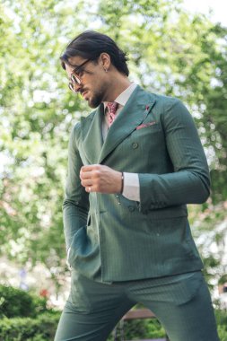 Yeşil takım elbiseli, güneş gözlüklü, yeşil yaprakların önünde el ele tutuşan seksi, zarif bir adam.