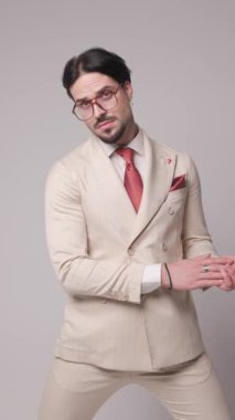 Havalı moda işadamı bej takım elbiseli, avuçlarını ovuyor, ceketi ayarlıyor, kollarını kavuşturuyor ve gri arka planda küstahça poz veriyor.