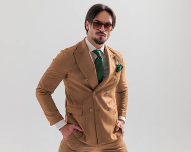 Gri arka planda el ele tutuşan kahverengi göğüslü, güneş gözlüklü şık moda adamı.