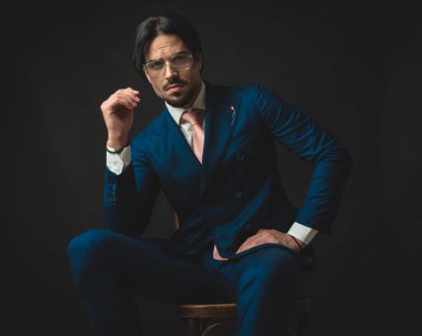 Modern iş adamı gözlüklü, klasik çift göğüslü lacivert takım elbiseli, dirseklerini kalçasında tutan, ileriye bakan ve oturan 