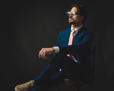 Gözlüklü, düşünceli, genç bir iş adamı. Bacak bacak üstüne atmış, dizleri üstüne çökmüş, siyah arka planda oturuyor.