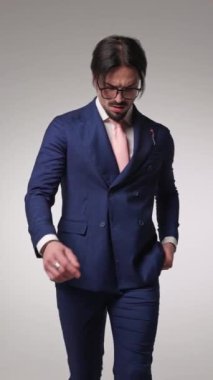 Lacivert takımlı seksi genç adam kravatını düzeltiyor ve gri arka planda el ele hareket ediyor.