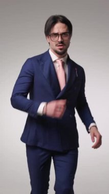 Lacivert takım elbiseli, kendine güvenen genç bir işadamı. Gözlüklü ve aceleyle ofise gidiyor. El kol hareketi yapıyor ve gri arka plana davet ediyor.