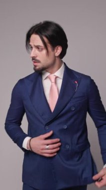 Havalı modacı taraf arıyor ve lacivert takımı ayarlıyor, el ele tutuşup, gri arka planda parmaklarını şıklatıyor.