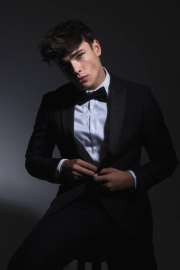 Maço, zarif bir adam siyah smokinin düğmelerini iliklerken karanlık arka planda oturuyor.