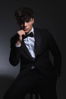 Siyah smokinli, şehvetli, zarif bir adam. El ele tutuşup, koyu gri arka planda otururken yüzüne dokunuyor.