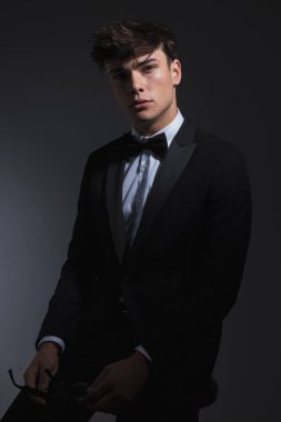 Siyah smokinli zarif bir genç adam elinde güneş gözlüğü tutarak koyu gri arka planda poz veriyor.