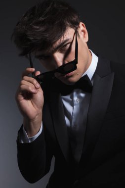 Siyah smokinli seksi genç damat koyu gri arka planda güneş gözlüğü takıyor ve ona bakıyor.