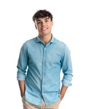 Mavi kot gömlekli mutlu genç adam, Çin pantolonunun cebinde el ele tutuşuyor, kameraya bakıyor ve beyaz arka planda dururken büyük bir gülümseme sergiliyor.