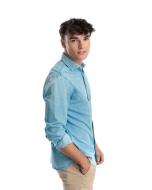 Mavi kotlu tişörtlü yakışıklı bir adam, Çin pantolonunun cebinde el ele tutuşuyor, kameraya bakıyor ve beyaz arka planda havalı poz veriyor.