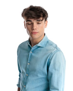 Stüdyoda beyaz arkaplan önünde dikilirken gözleri kapalı poz veren mavi kot gömlekli yorgun bir adam.