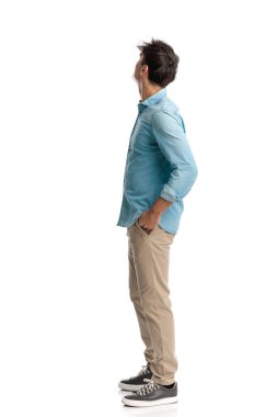 Mavi kotlu gömlekli ve elleri cebinde olan beyaz arka planda merak içinde genç bir öğrencinin yan görünüşü.