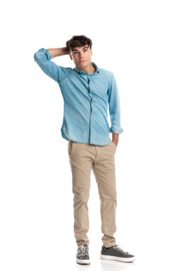 Mavi kot gömlekli kafası karışmış genç bir öğrenci. Elleri kafasının arkasını tırmalıyor. Cebinde ve beyaz arka planda duruyor.