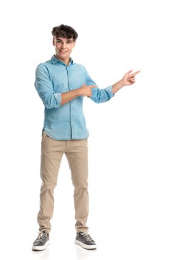 20 'li yaşlarda mutlu genç bir adam beyaz arka planda poz verirken parmaklarını yana doğru gösteriyor ve gülümsüyor.