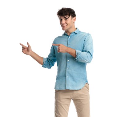 20 'li yaşlardaki mutlu öğrenci mavi kot gömlekli, parmaklarını yana doğru gösteren ve beyaz arka planda gülümseyen.