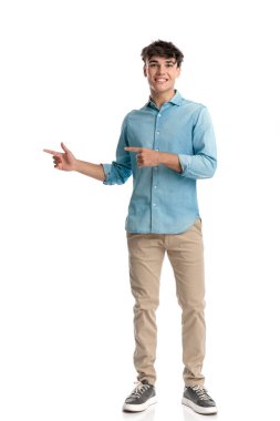 Mavi kot gömlekli sevimli genç öğrenci mutlu ve gülümserken beyaz arka planı işaret ediyor.