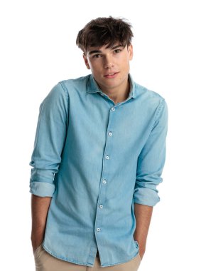 20 'li yaşlardaki ürkek genç bir öğrenci ceplerinde el ele tutuşup gülümsüyor, mavi kot gömlek giyiyor ve beyaz arka planda poz veriyor.