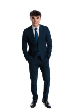 Lacivert takım elbiseli mutlu bir iş adamı ceplerinde el ele tutuşup beyaz arka planda gülümsüyor.