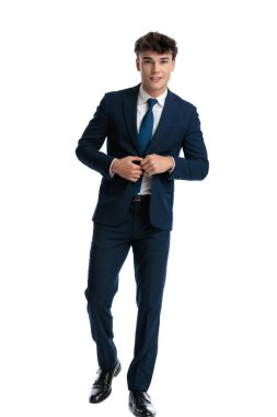 Kendine güvenen genç işletme öğrencisi lacivert takımını ilikliyor, beyaz arka planda yürüyor ve gülümsüyor.