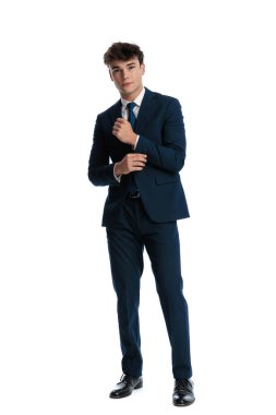 Takım elbise kollarını düzeltirken gülümseyen ve beyaz arka planın önünde duran çekici genç bir işletme öğrencisi.