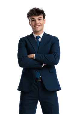 Stüdyoda beyaz arkaplanda poz verirken kollarını kavuşturup gülümseyen lacivert takım elbiseli mutlu genç bir işletme öğrencisi.