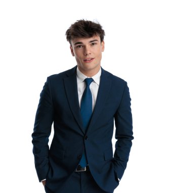 Lacivert takım elbiseli genç bir işletme öğrencisi ceplerinde el ele tutuşuyor ve stüdyodaki beyaz arka plana gülümsüyor.