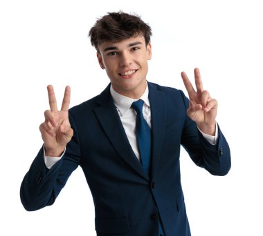 Lacivert takımlı mutlu genç öğrenci parmaklarıyla barış işareti yapıyor, gülümsüyor ve stüdyoda beyaz arkaplan önünde poz veriyor.