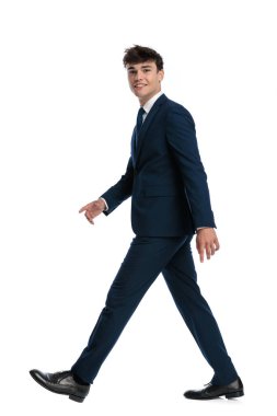 Mavili lacivert takım elbiseli zarif bir genç beyaz arka planda yürüyor ve gülümsüyor.