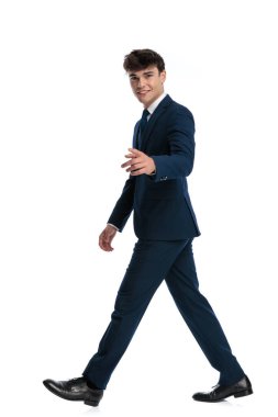 Mavili lacivert takım elbiseli, beyaz arka planda yürürken gülümseyen ve parmağını öne doğru gösteren kendinden emin genç adamın yan görüntüsü.