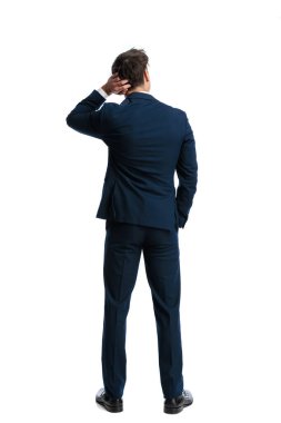 Kafasını kaşıyan ve elini cebine sokup beyaz arka planda gezen şaşkın, zarif iş adamının arka görüntüsü.