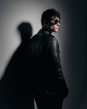 Güneş gözlüklü, elleri ceplerinde gri bir duvara yaslanmış ve yan tarafa bakan seksi genç oyuncunun arka görünüşü. 