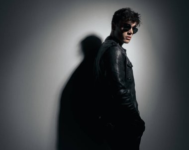 Siyah deri ceketli, güneş gözlüklü, elleri ceplerinde gri arka planda havalı bir modacının yan görüntüsü. 
