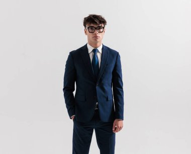 20 'li yaşlarda, lacivert takım elbiseli zarif genç bir işletme öğrencisi. Gri arka planda elleri ceplerinde poz veriyor.