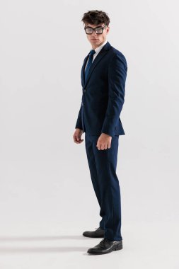 Mavili lacivert takımlı zarif bir genç gözlüklü ve gri arka planda duruyor.