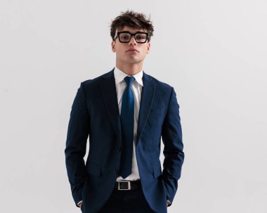 Lacivert takım elbiseli, kendine güvenen genç bir işadamı. Ceplerinde el ele tutuşup gri arka planda poz veriyor.