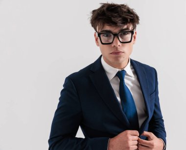 Gözlüklü genç iş adamının portresi lacivert takımı ayarlıyor ve gri arka planda poz veriyor.