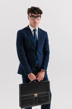 20 'li yaşlarda, lacivert takım elbiseli, bavul tutan ve gri arka planda duran seksi ve zarif bir adam.