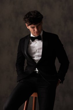 Siyah smokinli zarif bir genç adam elleri ceplerinde gözleri kapalı bej desenli arka planda otururken ışıklar gözlerini kör ediyor.