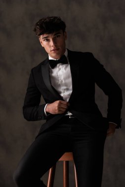 Siyah smokinini ayarlayan ve bej desenli arka planda otururken el ele tutuşan yirmili yaşlardaki çekici damadı.