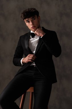 Siyah smokinli zarif genç adam papyon ve smokin ayarlıyor ve bej desenli arka planda tahta koltukta otururken poz veriyor.