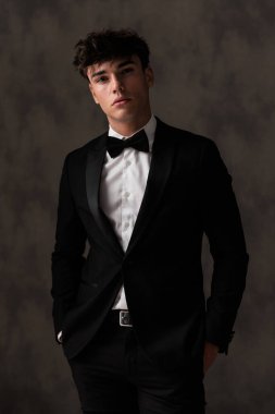 Siyah smokinli zarif genç damat el ele tutuşup bej desenli arka planda duruyor.