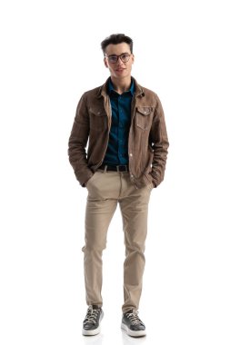 Kahverengi deri ceket ve gözlük giyerek beyaz arka planda ceplerini tutarken rahat, genç ve rahat bir manken.