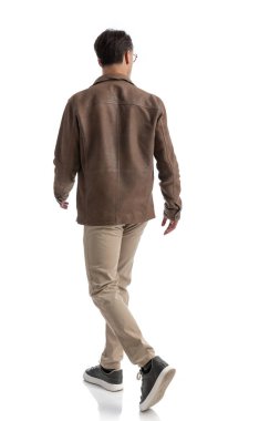 Kahverengi deri ceketli ve gözlüklü, beyaz arka planda yürüyen, tüm vücudunu gösteren zeki bir adamın arkadan görünüşü.