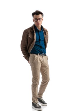 Deri ceket ve gözlük giymiş, beyaz arka planda dururken elleri arkada poz veren baştan çıkarıcı, zeki, sıradan bir adam.