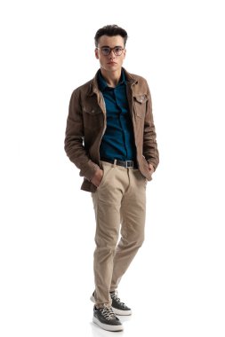 Deri ceketli ve gözlüklü genç adam ceplerini tutarken beyaz arka planda duruyor.