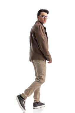 Deri ceket giyen, beyaz arka planda tek başına yürüyen akıllı, sıradan bir adamın yan görüntüsü.